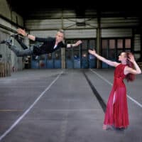 Magician performing a mid-air illusion with a female assistant in an industrial setting.